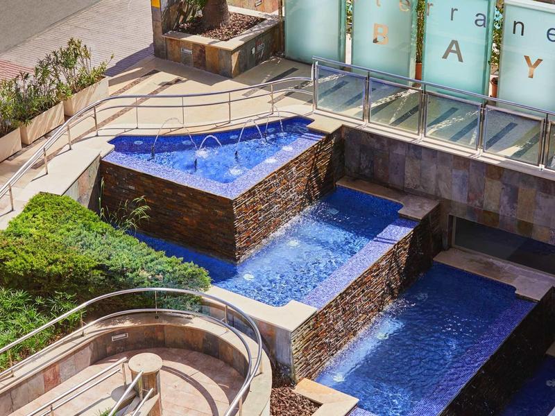 Dreistufiger blauer Wasserspringbrunnen aus Stein mit umliegenden Grünflächen und Treppen.