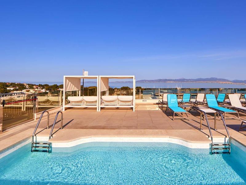 Klares blaues Schwimmbecken mit Liegestühlen und Doppelbetten unter blauem Himmel am Rand einer Terrasse.