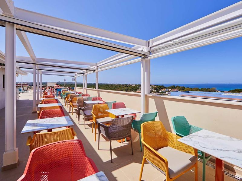Dachterrasse mit bunten Stühlen und Tischen, blauem Himmel und Meerblick im Hintergrund.
