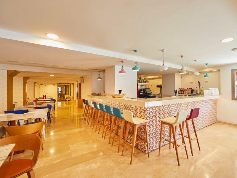 Heller, moderner Gastraum mit großem Barbereich und bunten Barhockern, warmes Licht und Holzboden.