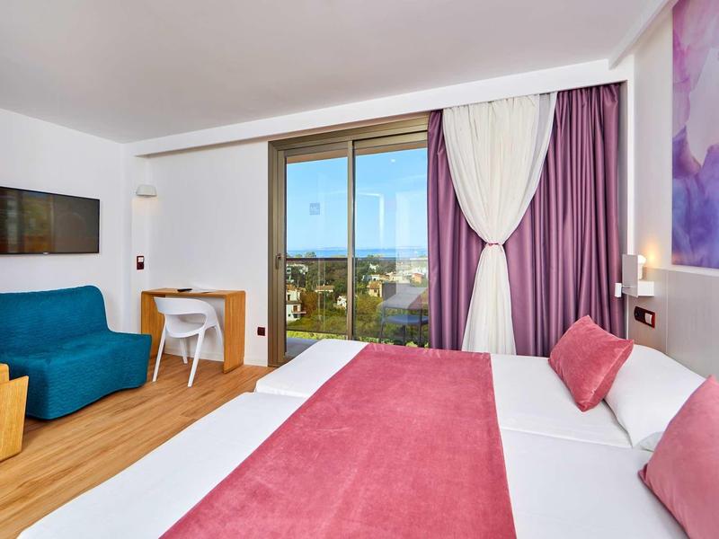 Helles Hotelzimmer mit großem Bett, lila Vorhängen, blauem Sessel, Schreibtisch und Stadtblick.