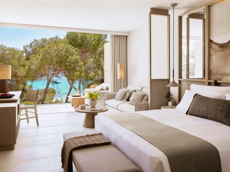 Jasny pokój hotelowy z dużym łóżkiem, sofą i widokiem na morze przez duże okna.