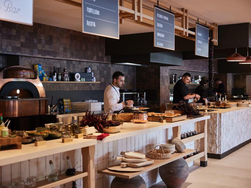 Strefa bufetu z drewnianymi ladami, różnymi potrawami i dwiema osobami przygotowującymi jedzenie.