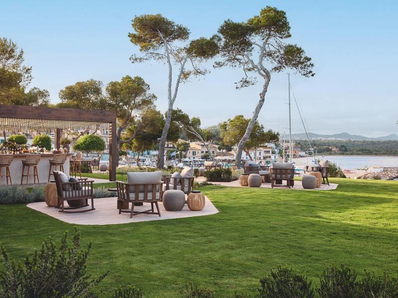 Zewnętrzny obszar wypoczynkowy z krzesłami, stolikami i widokiem na wodę i drzewa w słoneczny dzień.