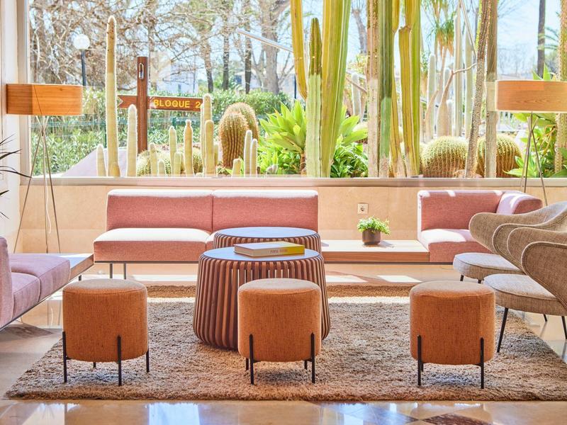 Bright living room with pink sofa, chairs, stools, and plants in front of large window.