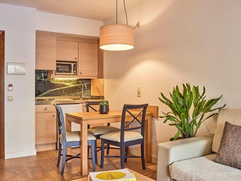 Cozy dining area with wooden table and chairs next to a small kitchenette under warm lighting.