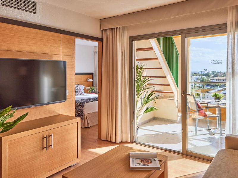 Bright hotel room with TV, sofa, balcony, exterior stairs, and city view.