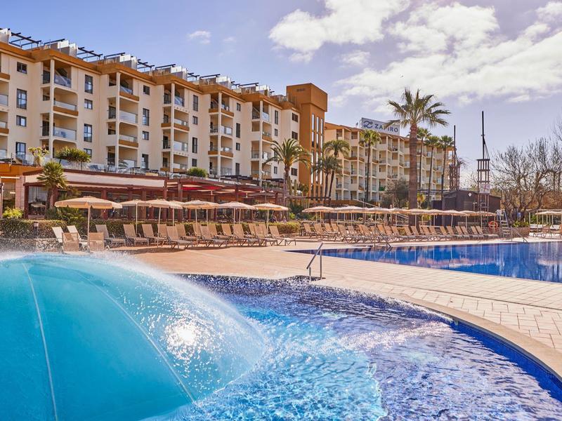 Large hotel with outdoor pool and lounge chairs under clear sky.