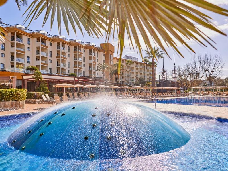 Blue pool with water feature in front of a multi-story hotel under palm trees.