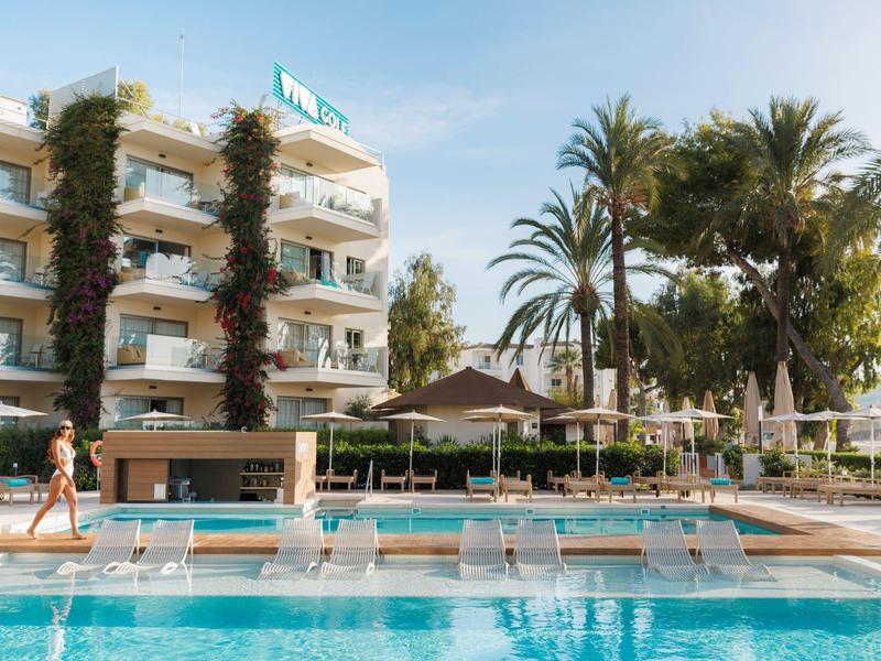 Piscine extérieure avec chaises longues devant un hôtel avec des palmiers sous un ciel clair.