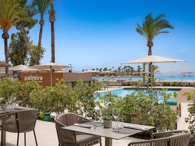 Terrasse avec tables dressées, vue sur piscine, palmiers et mer sous ciel clair.