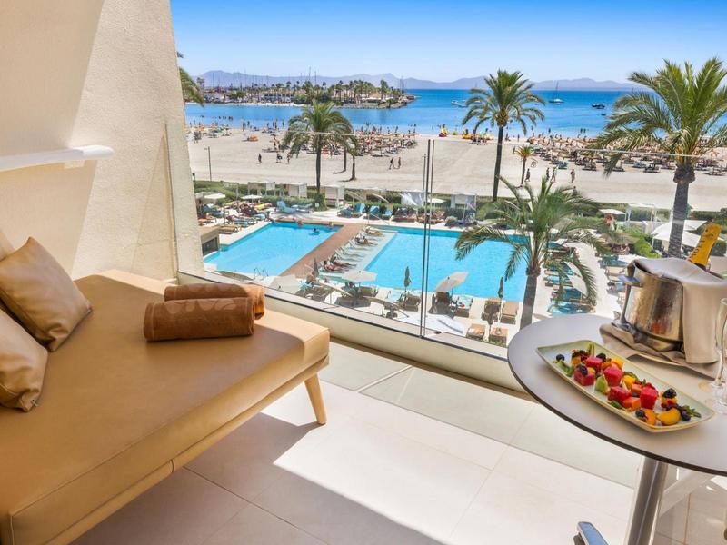 Balcon avec assise confortable et table avec champagne, vue sur piscine, palmiers et mer.