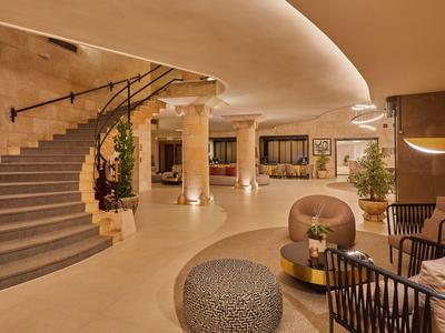 Hall d'hôtel spacieux avec escalier courbe, coin salon, plantes et éclairage chaleureux.