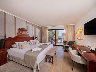 Habitación de hotel luminosa con cama doble, escritorio, TV y balcón con vista a la vegetación.