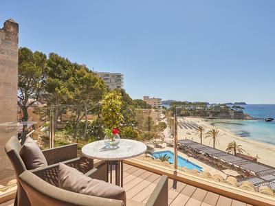 Balkon mit Sitzgruppe, Blick auf Strand, Meer, Bäume und Pool, klarer Himmel, helle Holzoberflächen.
