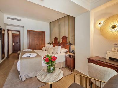 Chambre d'hôtel confortable avec un lit double, une tête de lit en bois, un bureau, un fauteuil et des fleurs fraîches sur une table ronde.