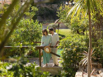 Couple marchant sur un petit pont en bois entouré d'une végétation luxuriante dans un jardin ensoleillé.
