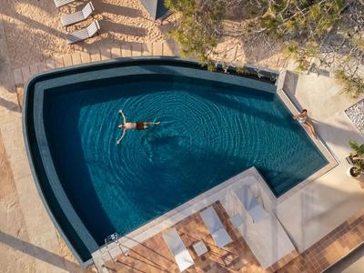 Vista aérea de una piscina abierta con una persona relajándose, rodeada de tumbonas y plantas.