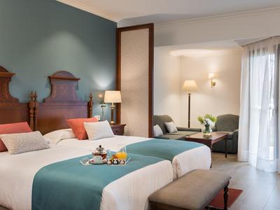 Chambre d'hôtel avec deux lits, accents turquoise, têtes de lit en bois et coin salon près de la fenêtre.