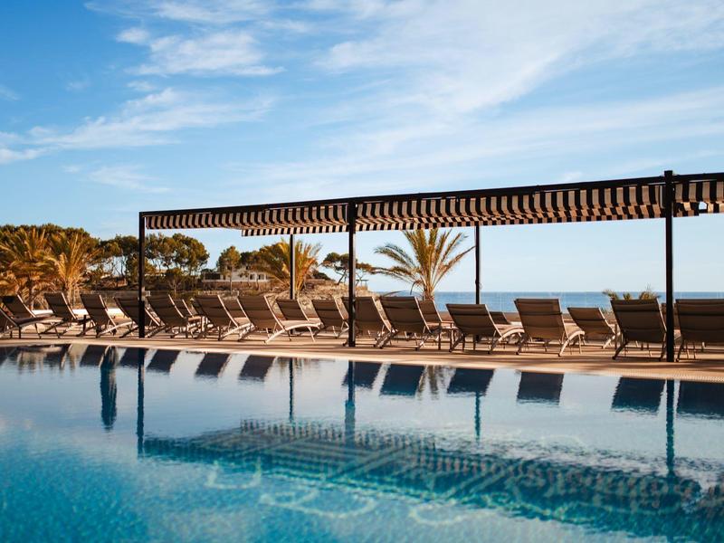 Alignement de chaises longues vides au bord d'une piscine sous une pergola avec des palmiers et ciel bleu.