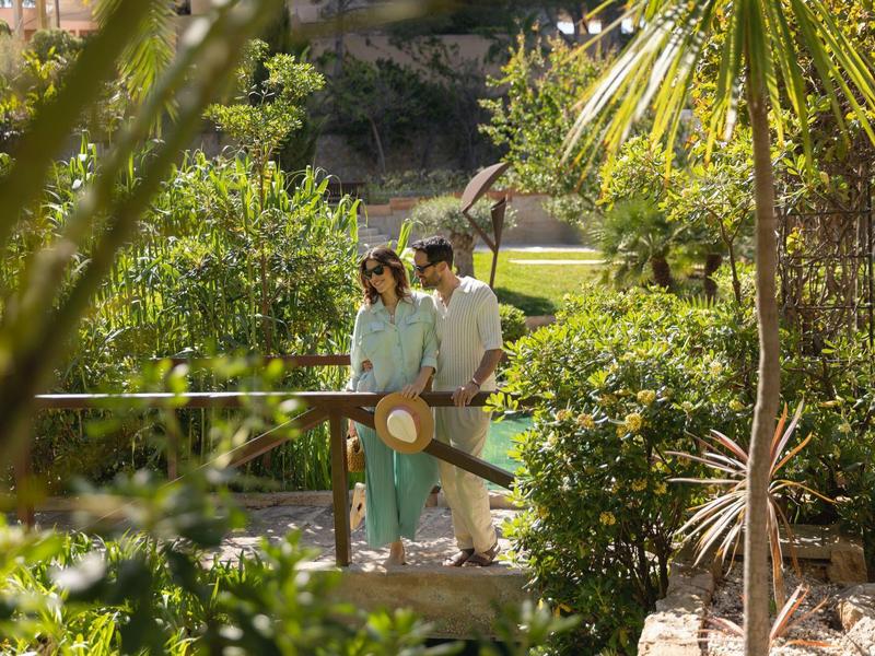 Couple marchant sur un petit pont en bois entouré d'une végétation luxuriante dans un jardin ensoleillé.