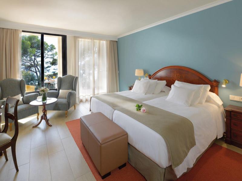 Chambre d'hôtel confortable avec grand lit, mur bleu, bureau et balcon vue sur les arbres.