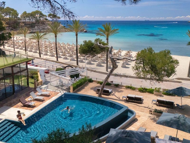 Station avec piscine, palmiers et transats surplombant une plage bleue claire.