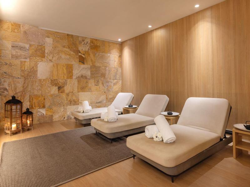 Salle de spa relaxante avec trois chaises longues, serviettes, murs en pierre et bois, éclairage chaleureux.