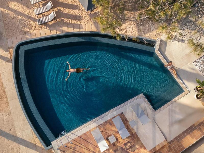Vue aérienne d'une personne flottant dans une piscine bleue de forme unique entourée de chaises longues et d'arbres.