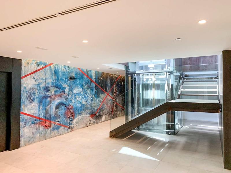 Moderní hotelová chodba s abstraktním nástěnným uměním, skleněným výtahem a schodištěm s přirozeným světlem
