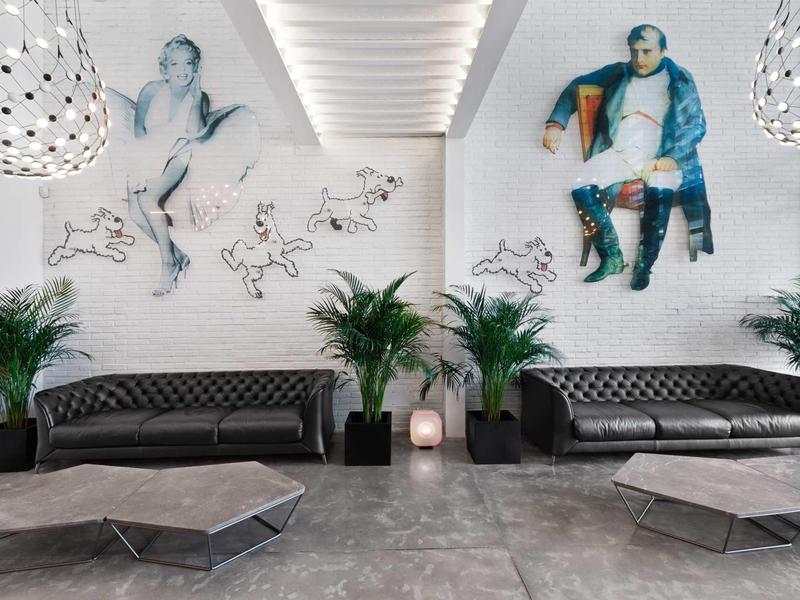 Moderní čekárna s černými pohovkami, rostlinami a nástěnným uměním v světlé místnosti.