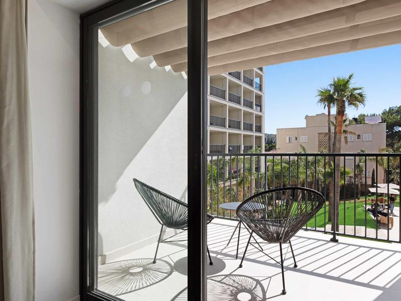 Balkon s dvěma židlemi a malým stolem s výhledem na hotelové budovy a palmy na slunci.