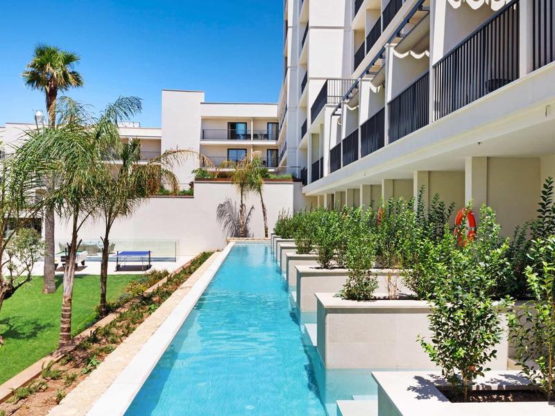 Moderní hotel s úzkým bazénem, palmami a upravenou zahradou pod jasnou oblohou.