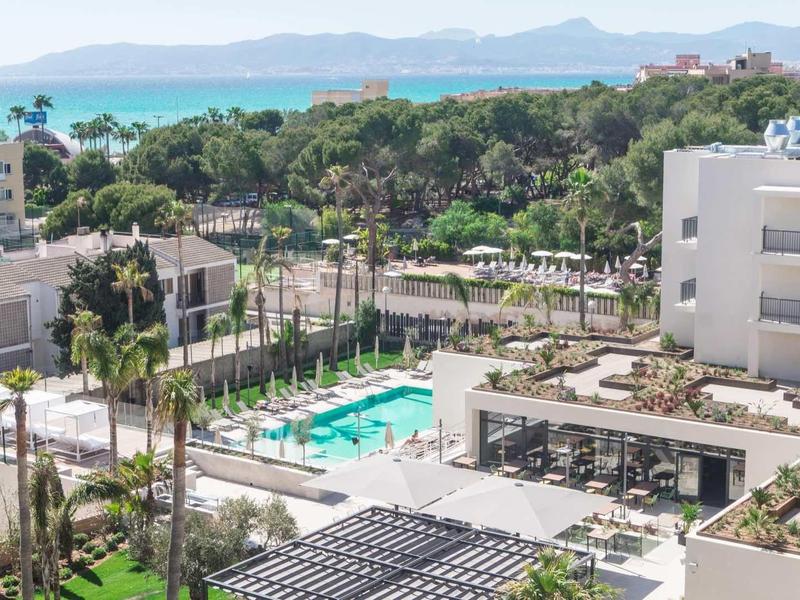 Moderní hotelový komplex s bazénem, zelení a výhledem na moře v pozadí.