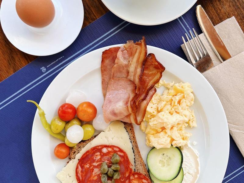 Talerz śniadaniowy z boczkiem, jajecznicą, pomidorami, ogórkiem, serem, bułką, kawą i jajkiem.