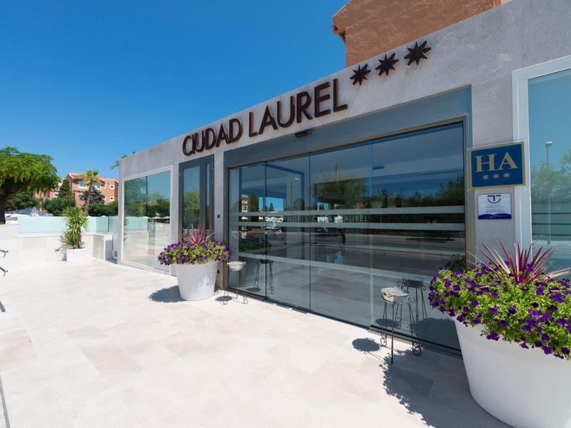 Nowoczesne wejście do hotelu ze szklaną elewacją i kwitnącymi roślinami pod błękitnym niebem