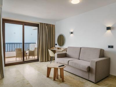 Zona de estar moderna con sofá gris, pequeña mesa de madera y acceso a balcón con vistas al mar.