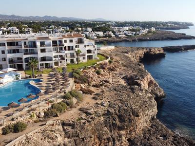Complejo hotelero con piscina en una costa rocosa con vistas al mar y montañas al fondo.