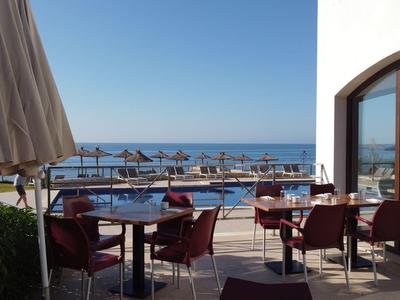 Terrassenbereich mit Tischen, Stühlen, Sonnenschirmen und Blick auf Pool und Meer bei klarem Himmel.