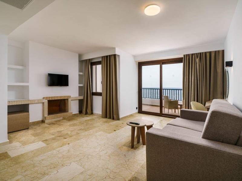 Sala luminosa con sofá, mesa pequeña, TV y acceso al balcón con vista al mar.