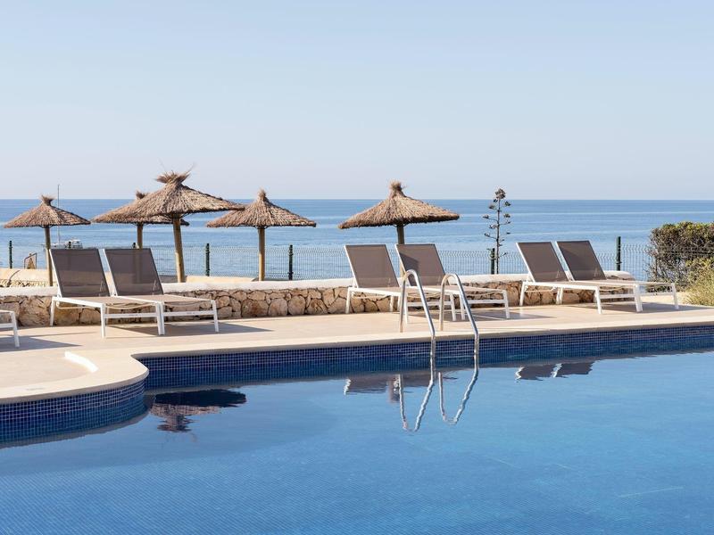 Piscina moderna con tumbonas y sombrillas de paja con vista al mar al fondo.