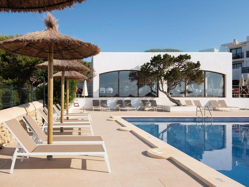 Zona de piscina con tumbonas y sombrillas frente a un edificio de hotel blanco y moderno.