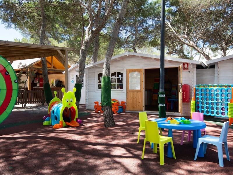 Área de juegos para niños colorida con mesas, sillas y juguetes bajo árboles.