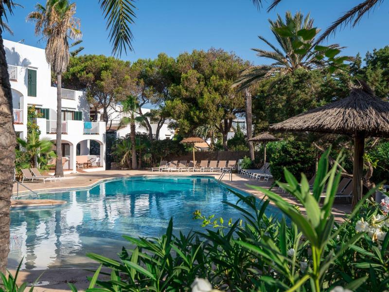 Piscina del hotel con sombrillas y tumbonas, rodeada de palmeras y vegetación verde.