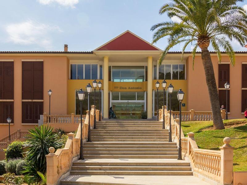 Entrée de bâtiment jaunâtre avec escaliers, palmiers et lampes sous un ciel clair.