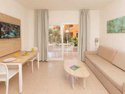 Helle Wohnzimmer mit beige Couch, kleinem Tisch, Fernseher, Schreibtisch und Zugang zu Terrasse mit Garten.