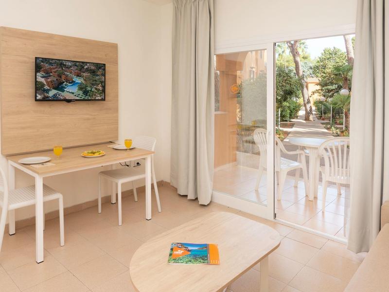 Salon lumineux avec table à manger, deux chaises blanches, télévision et vue sur terrasse avec jardin