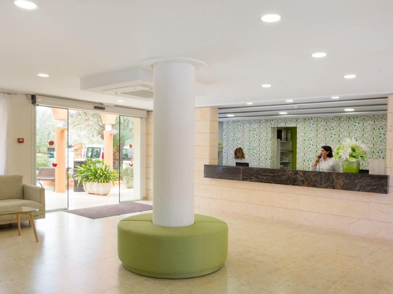 Hall d'hôtel lumineux avec réception, banquette verte circulaire autour d'un pilier et coin salon.