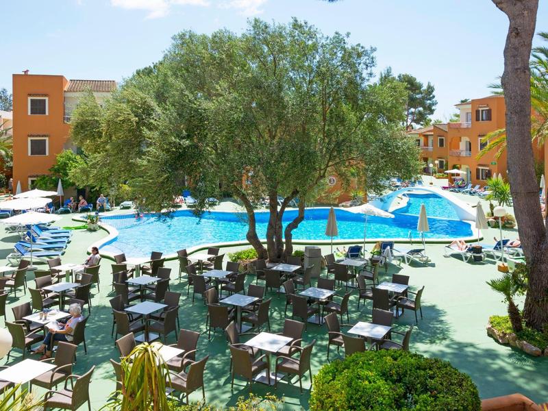 Grande piscine avec parasols et tables devant un hôtel avec des arbres et un ciel bleu.