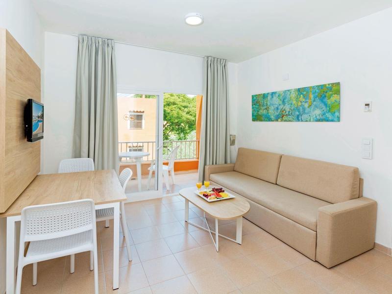 Salon moderne avec canapé, table basse, table à manger, télévision, couleurs claires et balcon donnant sur la verdure.